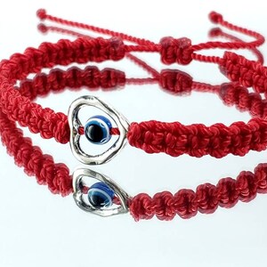 K&ouml;nnte beinhalten: Zwei rote Armb&auml;nder aus Schnur mit einem silbernen Herz-Anh&auml;nger mit einem blauen b&ouml;sen Auge. Die Armb&auml;nder sind mit einem Knoten zusammengebunden.