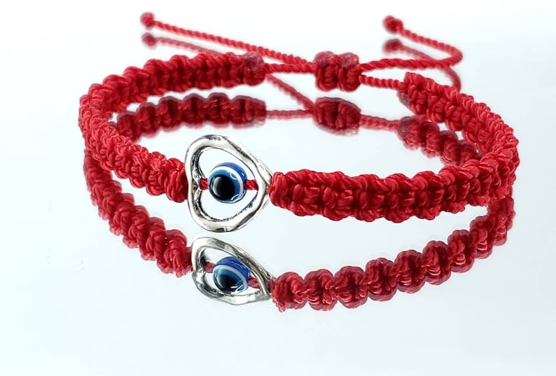 K&ouml;nnte beinhalten: Zwei rote Armb&auml;nder aus Schnur mit einem silbernen Herz-Anh&auml;nger mit einem blauen b&ouml;sen Auge. Die Armb&auml;nder sind mit einem Knoten zusammengebunden.