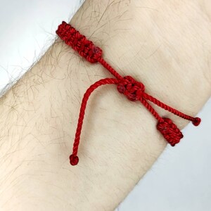 K&ouml;nnte beinhalten: Ein rotes Armband aus Schnur mit gekn&uuml;pften Enden. Das Armband ist um das Handgelenk einer Person gebunden.