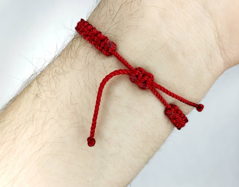 K&ouml;nnte beinhalten: Ein rotes Armband aus Schnur mit gekn&uuml;pften Enden. Das Armband ist um das Handgelenk einer Person gebunden.