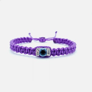 May include: A purple macrame bracelet with an adjustable sliding knot closure. The bracelet features a central charm with a blue and black evil eye design, framed by small, sparkling silver-colored accents. The bracelet is designed for wrist sizes up to 8 inches.