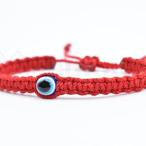 May include: Red string bracelet with a blue evil eye charm. The bracelet is knotted and has a sliding knot for adjusting the size.