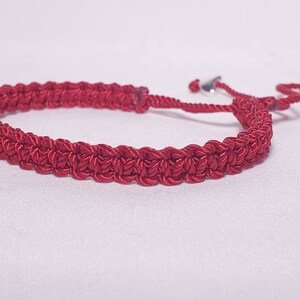 Puede incluir: Un brazalete trenzado rojo con detalles en metal plateado. El brazalete es ajustable con un nudo deslizante.