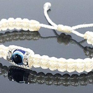 May include: Cream-colored macrame bracelet featuring a central blue evil eye bead with silver-colored accents. The bracelet is adjustable with a sliding knot closure. The woven design and the evil eye charm create a unique accessory.
