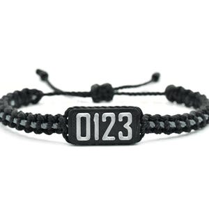 May include: A black braided cord bracelet featuring a central rectangular black plaque with the silver-colored numbers 0123 embossed on the front. The bracelet includes adjustable sliding knots and a thin gray cord woven through the black cord.