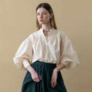 Op de afbeelding: Een crèmekleurige blouse met volumineuze mouwen en een hoge kraag, gecombineerd met een donkergroene rok. De blouse heeft een knoopsluiting aan de voorkant en de rok heeft een geplooide taille. Een ketting met een kruisvormige hanger is zichtbaar.