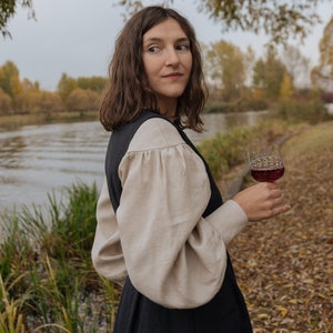 Op de afbeelding: Een vrouw in een zwarte jurk met een cr&egrave;mekleurige blouse en pofmouwen houdt een glas rode wijn vast. Ze staat in een veld met herfstbladeren en een rivier op de achtergrond.