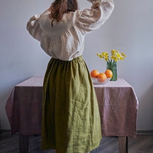 Pode incluir: Uma mulher vestindo uma blusa branca com mangas bufantes e uma saia de linho verde. Ela est&aacute; em p&eacute; em frente a uma mesa com um vaso de flores amarelas e uma tigela de laranjas.