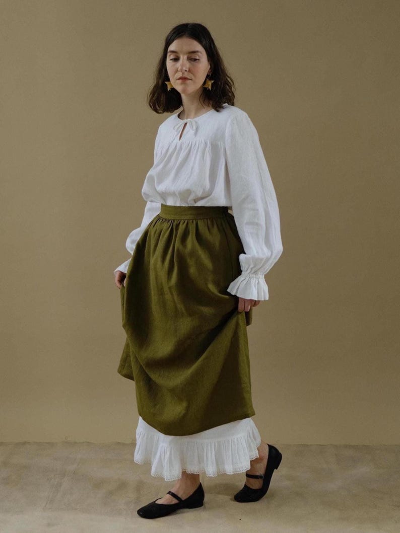 Pode incluir: Uma mulher vestindo uma blusa de linho branco com decote drapeado e mangas compridas, uma saia de linho verde escuro com renda branca e sapatos pretos. Ela est&aacute; usando brincos de estrela dourados.