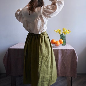 Op de afbeelding: Een vrouw draagt een wit linnen blouse met een groene linnen rok en witte sokken. Ze staat voor een tafel met een roze tafelkleed en een vaas met gele bloemen. Er staan sinaasappelen in een kom op de tafel.