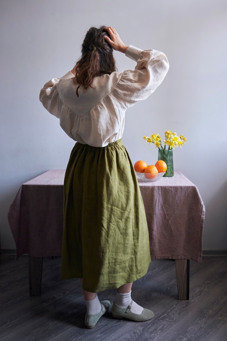 Op de afbeelding: Een vrouw draagt een wit linnen blouse met een groene linnen rok en witte sokken. Ze staat voor een tafel met een roze tafelkleed en een vaas met gele bloemen. Er staan sinaasappelen in een kom op de tafel.