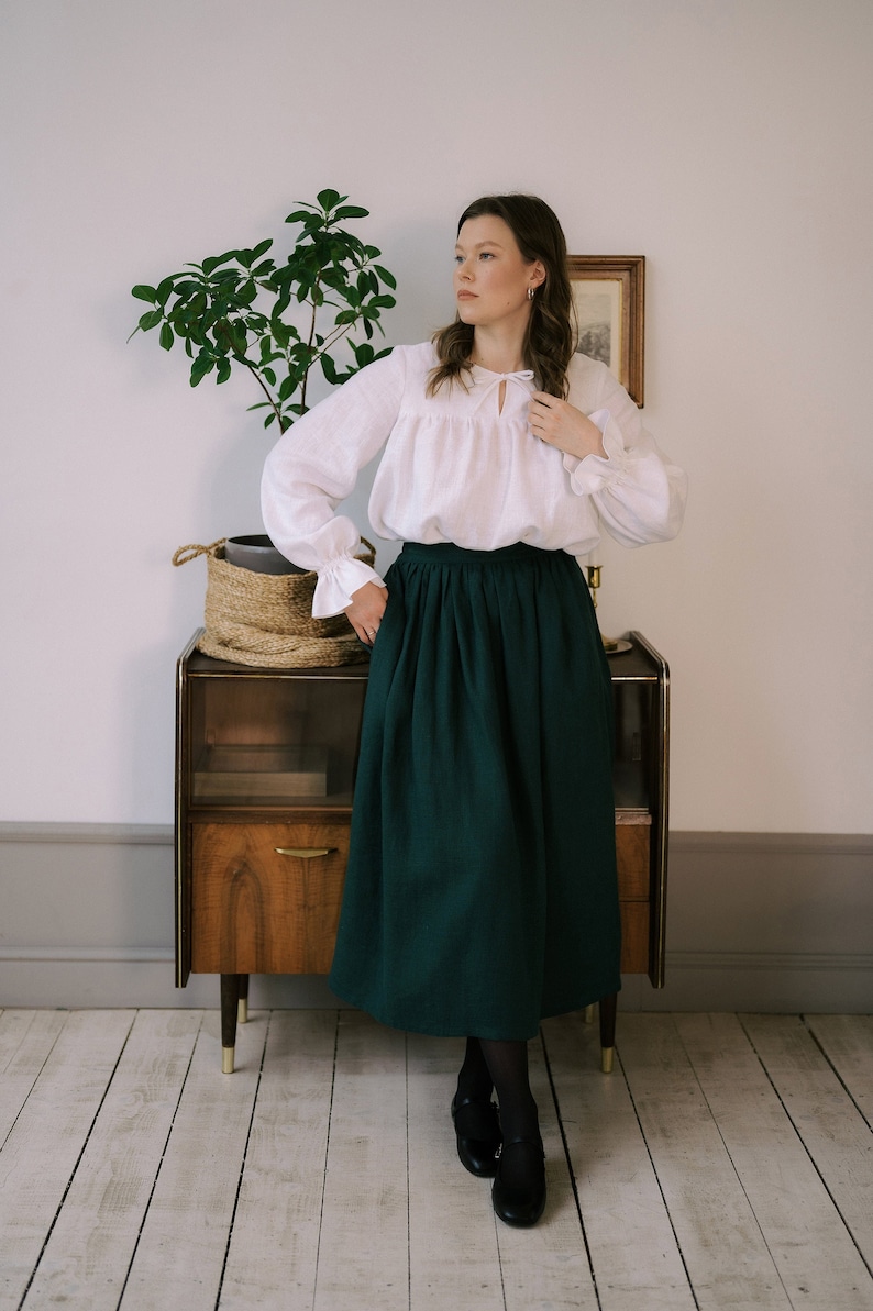Pode incluir: Uma mulher vestindo uma blusa de linho branca com um decote franzido e uma saia midi verde escura. Ela est&aacute; em p&eacute; em frente a um arm&aacute;rio de madeira com uma porta de vidro.