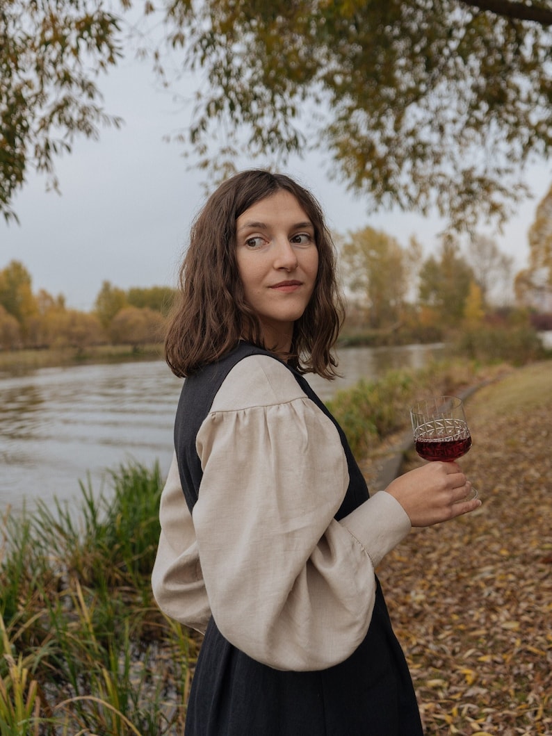 Op de afbeelding: Een vrouw in een zwarte jurk met een cr&egrave;mekleurige blouse en pofmouwen houdt een glas rode wijn vast. Ze staat in een veld met herfstbladeren en een rivier op de achtergrond.