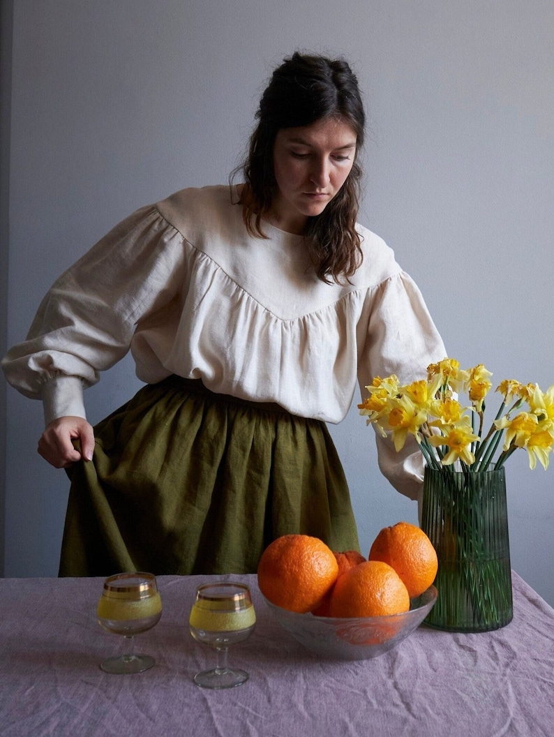 Op de afbeelding: Een vrouw in een wit blouse en een groene rok staat voor een tafel met een kom vol sinaasappelen en een vaas met gele narcissen.