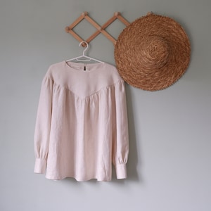 Op de afbeelding: Een licht beige linnen blouse met een ronde hals en lange mouwen hangt aan een houten hanger. De blouse heeft een geplooide detail aan de hals en een ontspannen pasvorm.