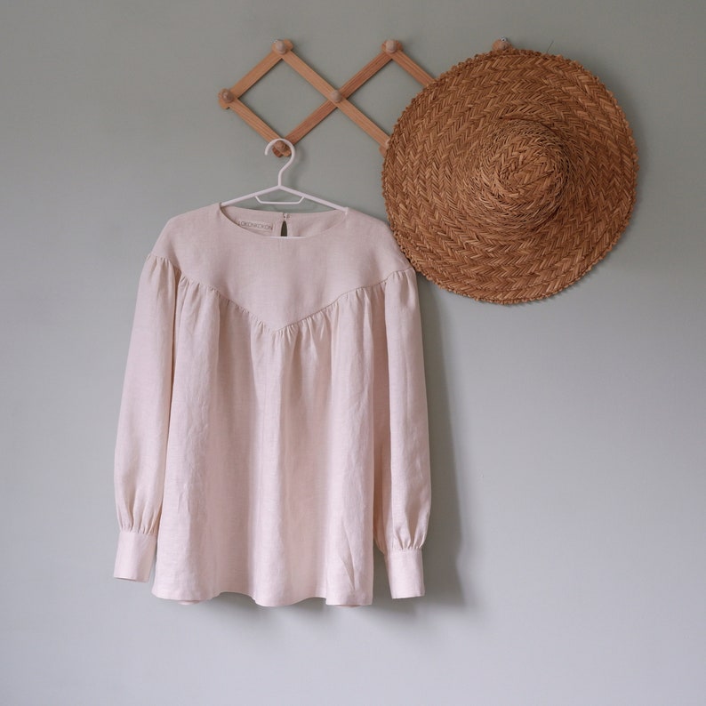 Op de afbeelding: Een licht beige linnen blouse met een ronde hals en lange mouwen hangt aan een houten hanger. De blouse heeft een geplooide detail aan de hals en een ontspannen pasvorm.