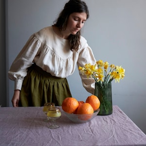 Op de afbeelding: Een vrouw in een wit blouse en een groene rok staat aan een tafel met een kom vol sinaasappelen en een vaas met gele narcissen.