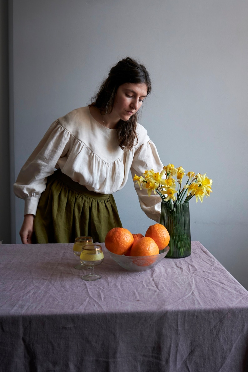 Op de afbeelding: Een vrouw in een wit blouse en een groene rok staat aan een tafel met een kom vol sinaasappelen en een vaas met gele narcissen.