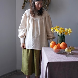 Op de afbeelding: Een vrouw in een wit linnen blouse en een groene linnen rok staat voor een tafel met een kom vol sinaasappelen en een vaas met gele narcissen. De tafel heeft een lichtpaarse tafelkleed.