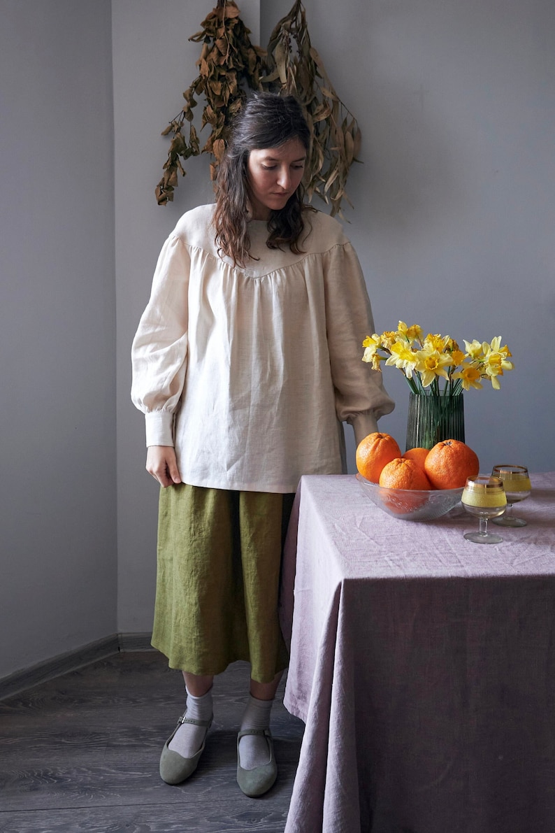 Op de afbeelding: Een vrouw in een wit linnen blouse en een groene linnen rok staat voor een tafel met een kom vol sinaasappelen en een vaas met gele narcissen. De tafel heeft een lichtpaarse tafelkleed.