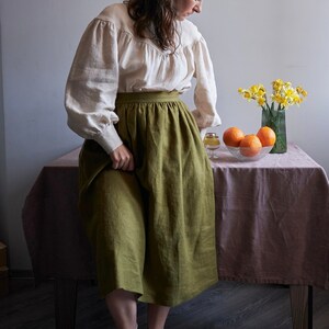 Op de afbeelding: Een vrouw in een wit blouse met een groene linnen rok en groene schoenen staat naast een tafel met een vaas met gele bloemen en een kom met sinaasappelen.