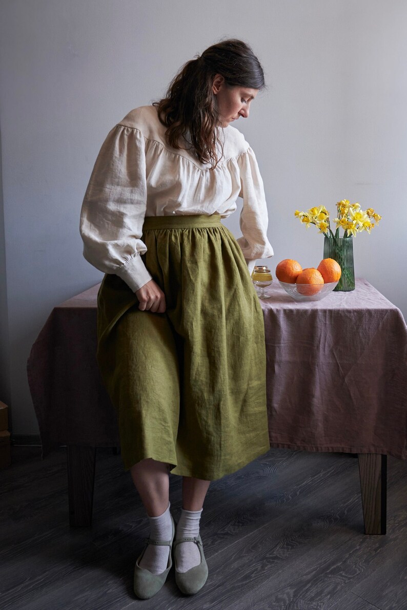 Op de afbeelding: Een vrouw in een wit blouse met een groene linnen rok en groene schoenen staat naast een tafel met een vaas met gele bloemen en een kom met sinaasappelen.