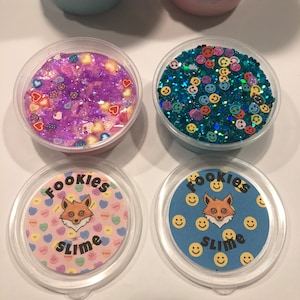 May include: Four containers of slime with different designs. Two containers have a fox face with the text "Fookies Slime" on the lid. One container has a blue glitter slime with smiley faces. The other container has a purple glitter slime with hearts.
