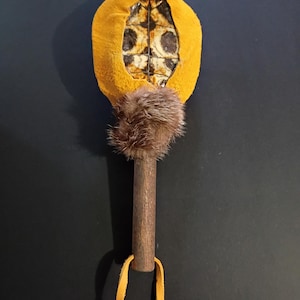May include: A handcrafted rattle with a wooden handle, a yellow suede-like top, and a fur accent. The rattle's top features a patterned design. A yellow leather strap hangs from the handle.
