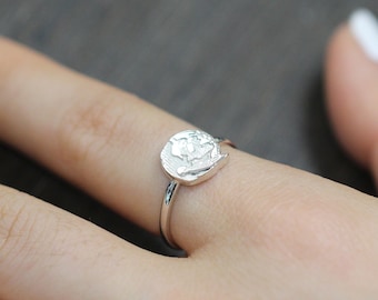 May include: A silver ring with a textured, round, silver design. The ring is on a person's finger.