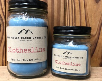 May include: Two glass candle jars with black lids. The larger jar is 16 ounces and has a burn time of 100-110 hours. The smaller jar is 8 ounces and has a burn time of 50-60 hours. Both jars have a blue candle inside and a brown label with white text that reads "Bear Creek Ranch Candle Co. Utopia, Texas Clothesline".