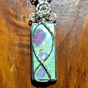 Puede incluir: Un colgante de alambre plateado con una piedra preciosa verde y rosa y una cadena plateada.