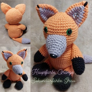 May include: A crocheted fox plush toy with orange fur, grey ears and nose, and black feet. The toy has green eyes and a black nose. The text "Hauptfarbe: Orange Sekund&auml;rfarbe: Grau" is written below the toy.