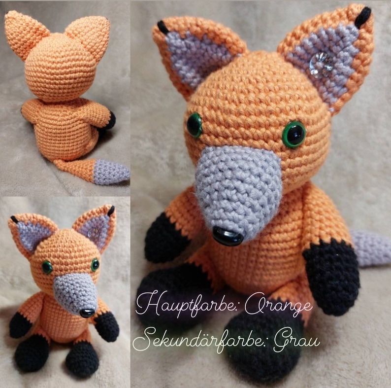 May include: A crocheted fox plush toy with orange fur, grey ears and nose, and black feet. The toy has green eyes and a black nose. The text "Hauptfarbe: Orange Sekund&auml;rfarbe: Grau" is written below the toy.