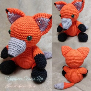 May include: A crocheted fox plush toy made with orange, grey and black yarn. The fox is sitting with its tail curled behind it. The text on the image reads "Hauptfarbe: Orange-Rot Sekund&auml;rfarbe: Grau".