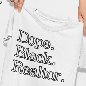 May include: White t-shirt with the text "Dope. Black. Realtor." in a bold, black font. The shirt is held up by a person's hands, showcasing the design. The upper left corner features a stylized logo.