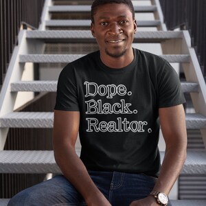 May include: Black t-shirt with the text "Dope. Black. Realtor." in a white, stylized font. The person in the image is wearing blue jeans and a watch with a brown leather band. The background is a metal staircase.