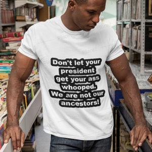 May include: White t-shirt with a black text design that reads "Don't let your president get your ass whooped. We are not our ancestors!" The shirt is worn by a person in a bookstore setting.