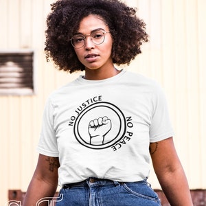 May include: White t-shirt with the text "NO JUSTICE NO PEACE" encircling a raised fist graphic. The shirt is paired with blue denim jeans. The image is set against a blurred yellow background.