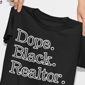 May include: Black t-shirt with the words "Dope. Black. Realtor." in white, stylized font. The shirt is held up by a person's hands. The upper left corner has a logo with the letters "S M T".