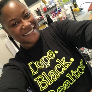 May include: Black long-sleeve shirt with the text "Dope. Black. Realtor" in a neon green font. The shirt is worn by a person smiling at the camera. A sign that says "create" is visible in the background.