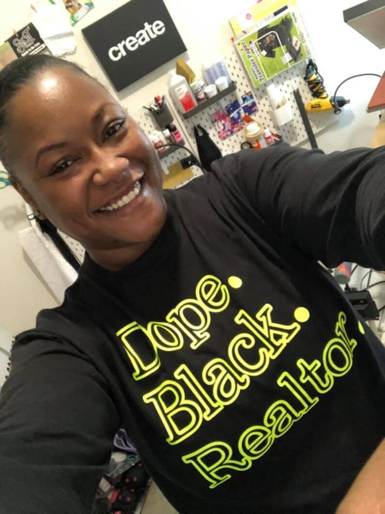 May include: Black long-sleeve shirt with the text "Dope. Black. Realtor" in a neon green font. The shirt is worn by a person smiling at the camera. A sign that says "create" is visible in the background.