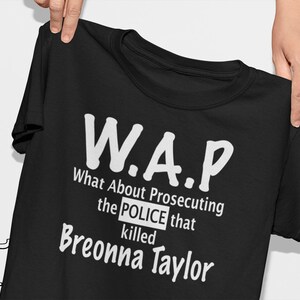 May include: Black t-shirt with white text. The text reads "W.A.P. What About Prosecuting the POLICE that killed Breonna Taylor." The shirt is held up by two hands.