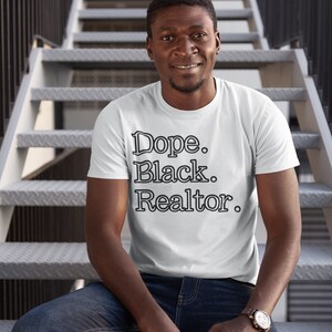 May include: White t-shirt with the text "Dope. Black. Realtor." in a black, bold font. The person is wearing blue jeans and a watch with a brown leather strap. The background is a metal staircase.