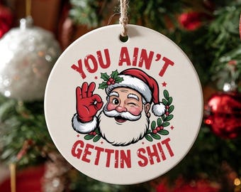 May include: Round Christmas ornament with a Santa Claus graphic. Santa is winking and giving the okay sign. The text on the ornament reads "YOU AIN'T GETTIN SHIT". The ornament is hanging on a Christmas tree.