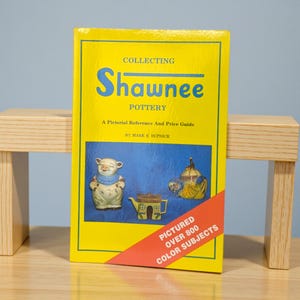 May include: A yellow book titled "Collecting Shawnee Pottery" with blue text and a red diagonal banner. The cover features images of Shawnee pottery pieces. The book is propped up on a small wooden stand.
