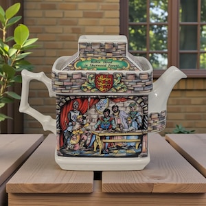 Puede incluir: Una tetera de cerámica con un diseño de castillo, que representa a Robin Hood en el bosque de Sherwood. La tetera tiene un asa y un pico blancos, con un patrón de ladrillos decorativo alrededor de la parte superior e inferior. El texto "Robin Hood in Sherwood Forest" es visible.