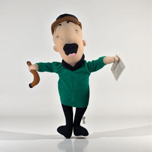 Puede incluir: Muñeco de peluche de un personaje de dibujos animados con cara bronceada, pelo negro y sombrero marrón. El muñeco lleva camisa verde, pantalón negro y zapatos negros. Sostiene un bastón marrón y una etiqueta.