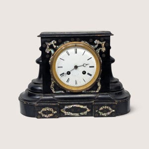 May include: A black mantel clock with a white circular face, black Roman numerals, and thin black hands. The clock features a gold-toned bezel and is decorated with inlaid iridescent mother-of-pearl pieces arranged in ornate patterns across the front.