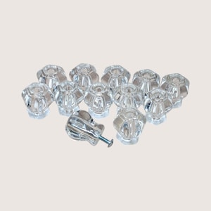 May include: A collection of clear, faceted glass cabinet knobs. The knobs have a flower-like design and are accompanied by a single knob with a screw attached. The knobs are arranged on a light background.