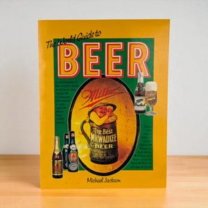 May include: A hardcover book titled "The World Guide to BEER" with a yellow cover. The cover features a large illustration of a beer mug with a man inside, and several beer bottles. The author's name, Michael Jackson, is at the bottom.
